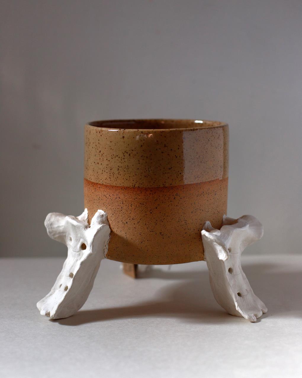 Handmade two-tone ceramic planter with a matte and gloss glaze, supported by three hand-sculpted ceramic replicas of deer or raccoon sacrum bones, displayed on a neutral background.