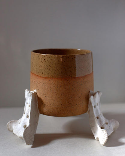 Handmade two-tone ceramic planter with a matte and gloss glaze, supported by three hand-sculpted ceramic replicas of deer or raccoon sacrum bones, displayed on a neutral background.